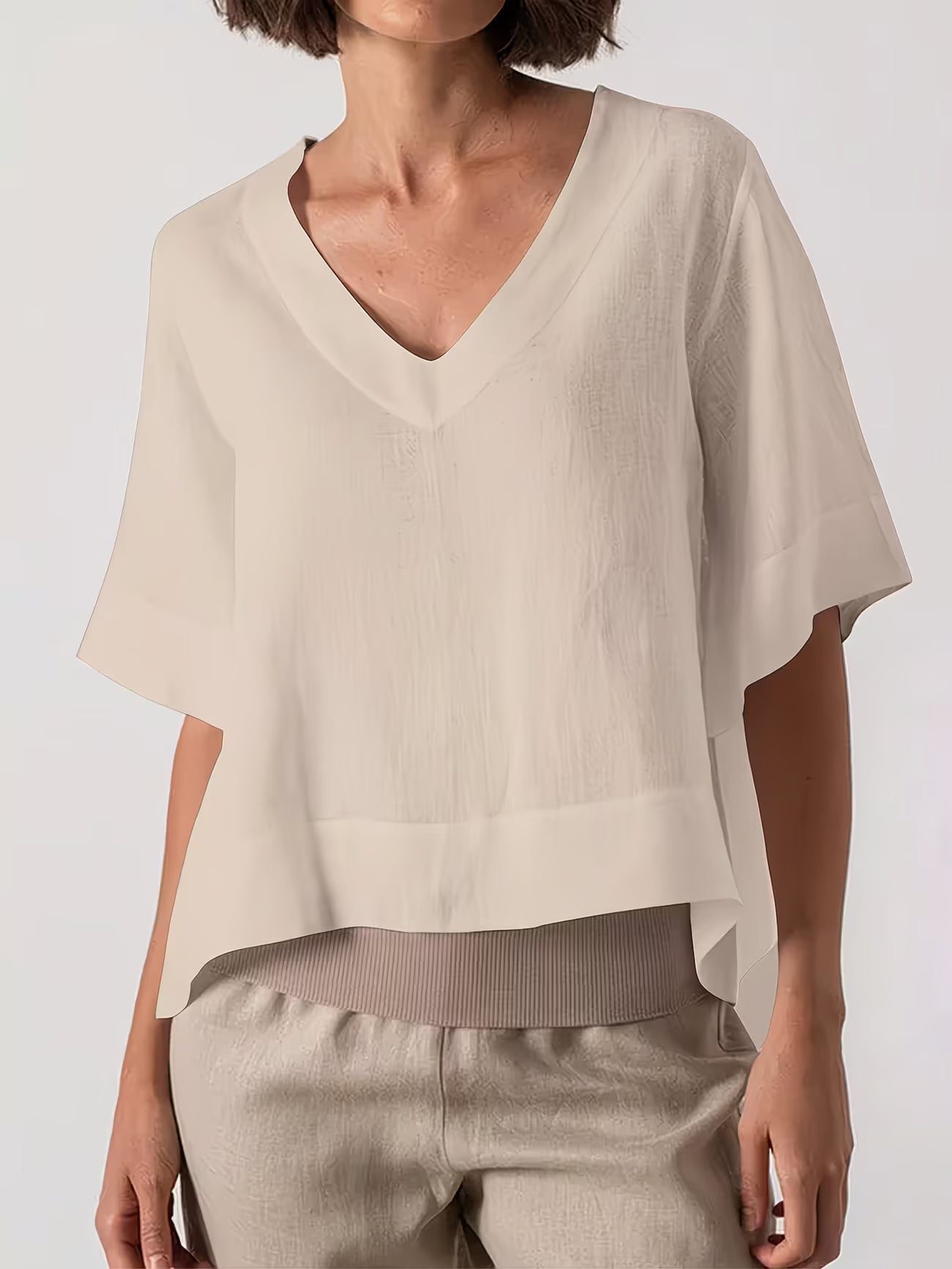 A woman wearing a light beige short-sleeve V-neck top with a relaxed, draped fit and light beige pants, photographed against a clean light gray studio background.