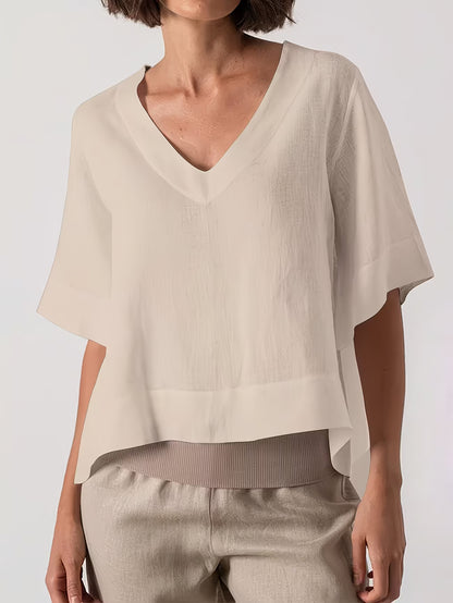 A woman wearing a light beige short-sleeve V-neck top with a relaxed, draped fit and light beige pants, photographed against a clean light gray studio background.