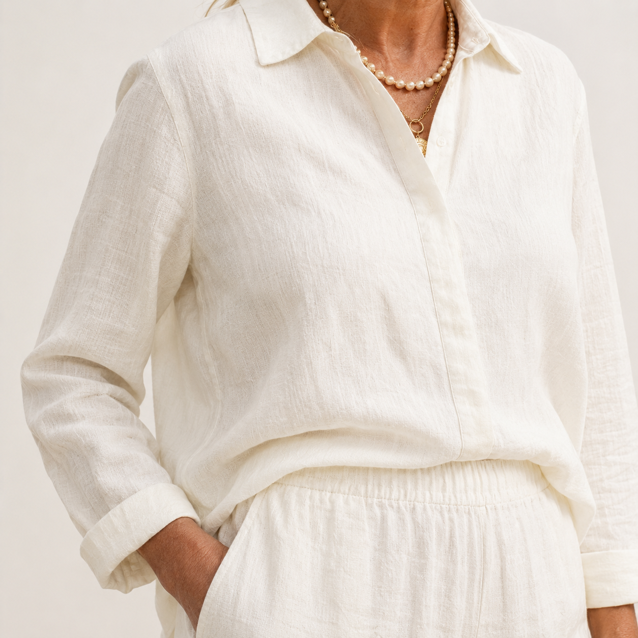 A close-up of a person wearing a loose white long-sleeve linen button-up shirt with a collar, paired with matching white bottoms and layered pearl necklaces.