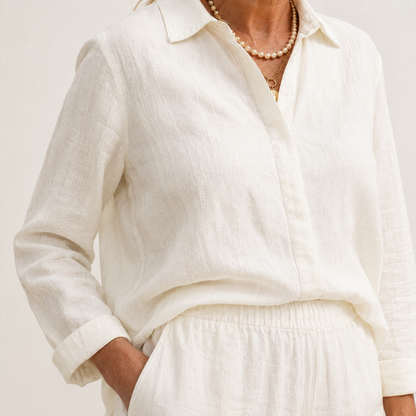 A close-up of a person wearing a loose white long-sleeve linen button-up shirt with a collar, paired with matching white bottoms and layered pearl necklaces.