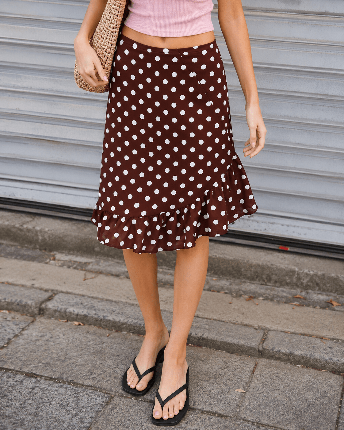 A woman wearing a brown midi skirt with white polka dots and a ruffled hem, paired with a pink crop top and black flip-flops, standing in front of a gray metal shutter.