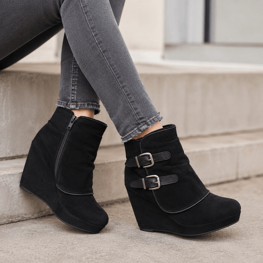 Black wedge ankle boots with dual buckle straps and a side zipper, shown on a model wearing gray jeans sitting on concrete steps.
