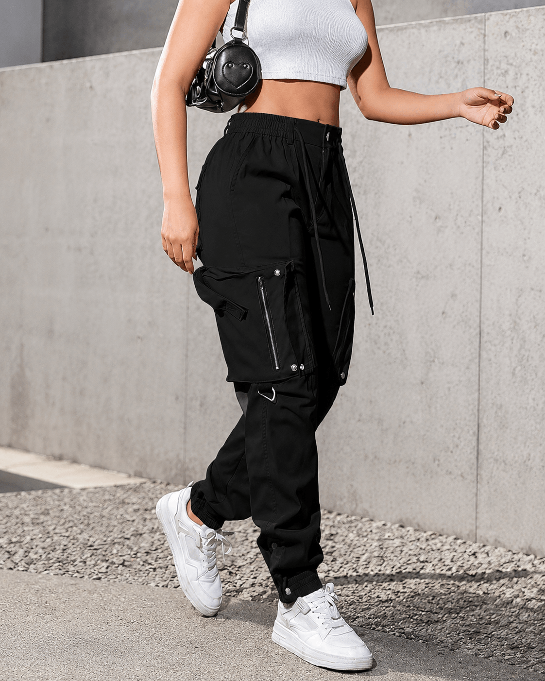 A person wearing black cargo jogger pants with white sneakers and a white crop top, standing outdoors against a gray concrete wall.