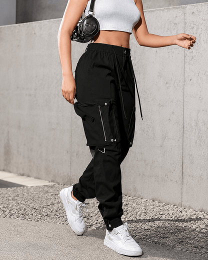 A person wearing black cargo jogger pants with white sneakers and a white crop top, standing outdoors against a gray concrete wall.