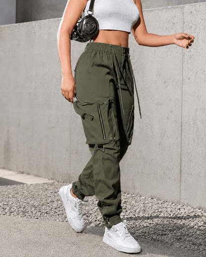 A person wearing olive green cargo jogger pants with side zippers and flap pockets, paired with a white crop top and white sneakers, standing outdoors against a concrete wall.