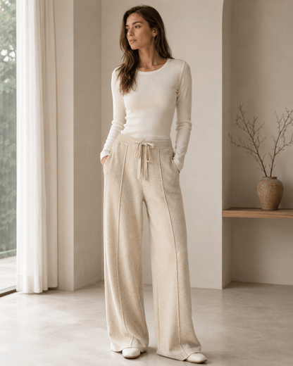 A woman stands in a bright, minimalist room wearing a cream long-sleeve knit top and matching cream wide-leg trousers with a drawstring waist.