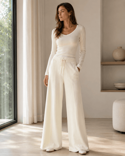 A woman stands in a bright, modern room wearing a cream-colored long-sleeve knit top and matching wide-leg trousers.