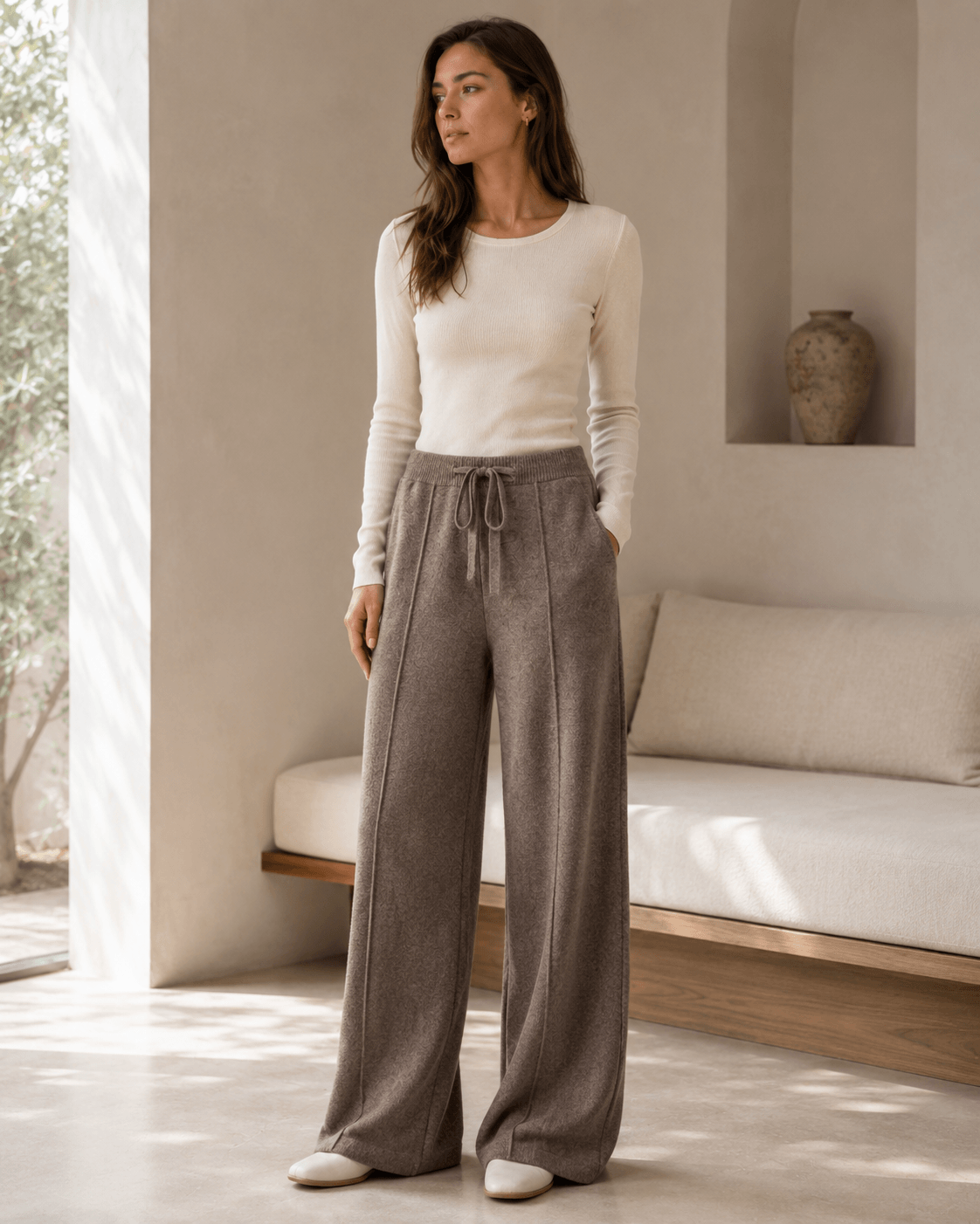 A woman stands in a bright, minimalist room wearing a fitted cream long-sleeve top and high-waisted, wide-leg taupe drawstring pants.