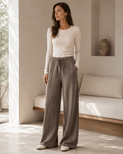 A woman stands in a bright, minimalist room wearing a fitted cream long-sleeve top and high-waisted, wide-leg taupe drawstring pants.