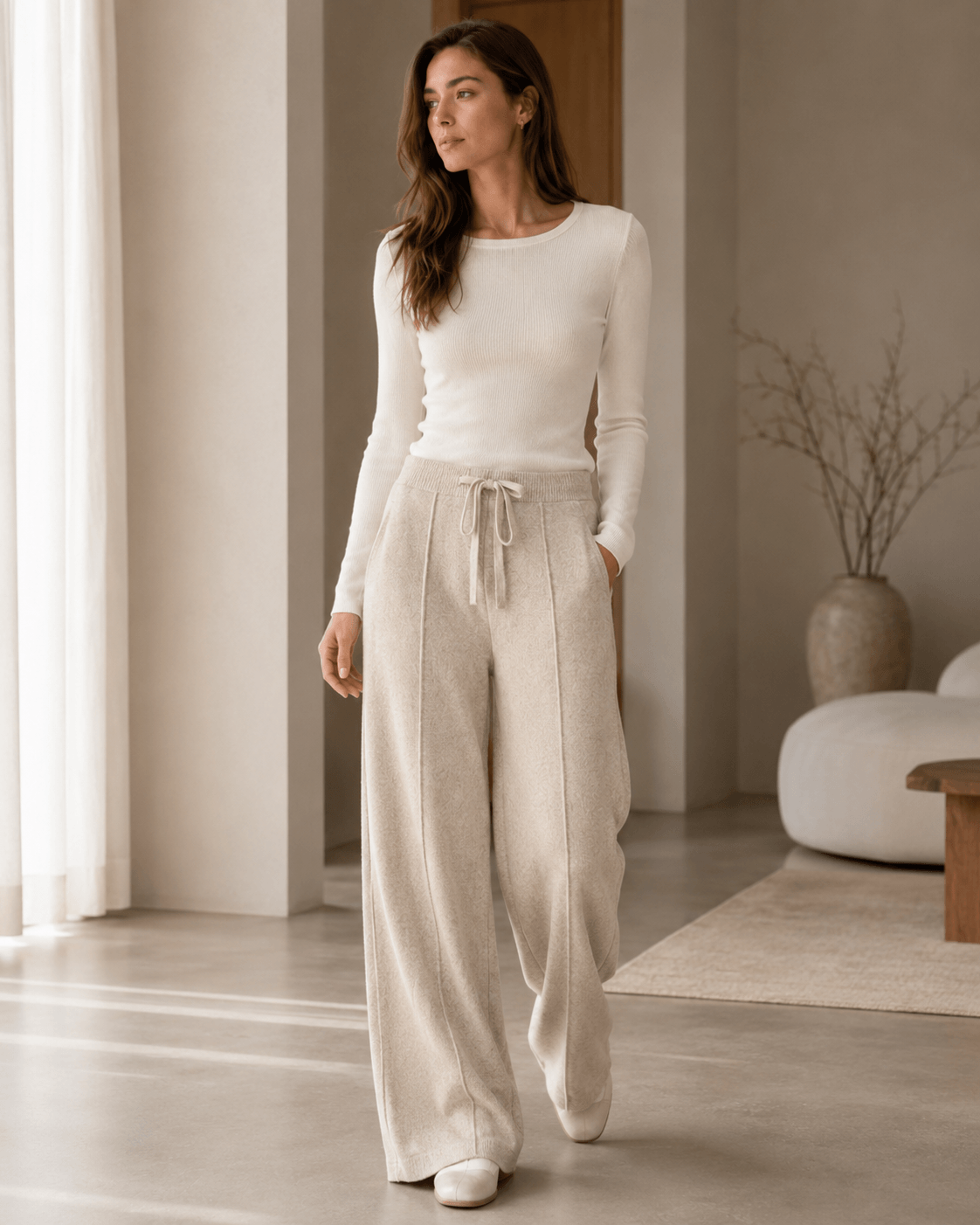 A woman wearing a cream long-sleeve knit top and matching cream wide-leg drawstring pants, standing in a bright, modern minimalist room.