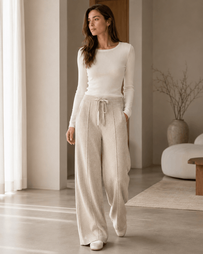 A woman wearing a cream long-sleeve knit top and matching cream wide-leg drawstring pants, standing in a bright, modern minimalist room.