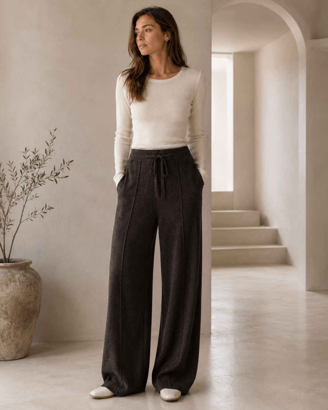 A woman stands in a minimalist interior wearing a fitted cream long-sleeve top and dark charcoal wide-leg drawstring pants, with a large vase and archway visible in the background.