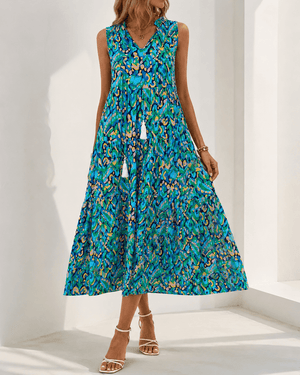 A woman wearing a sleeveless V-neck midi dress with a vibrant blue, green, and orange abstract floral print, paired with beige strappy high-heeled sandals in a bright white room.