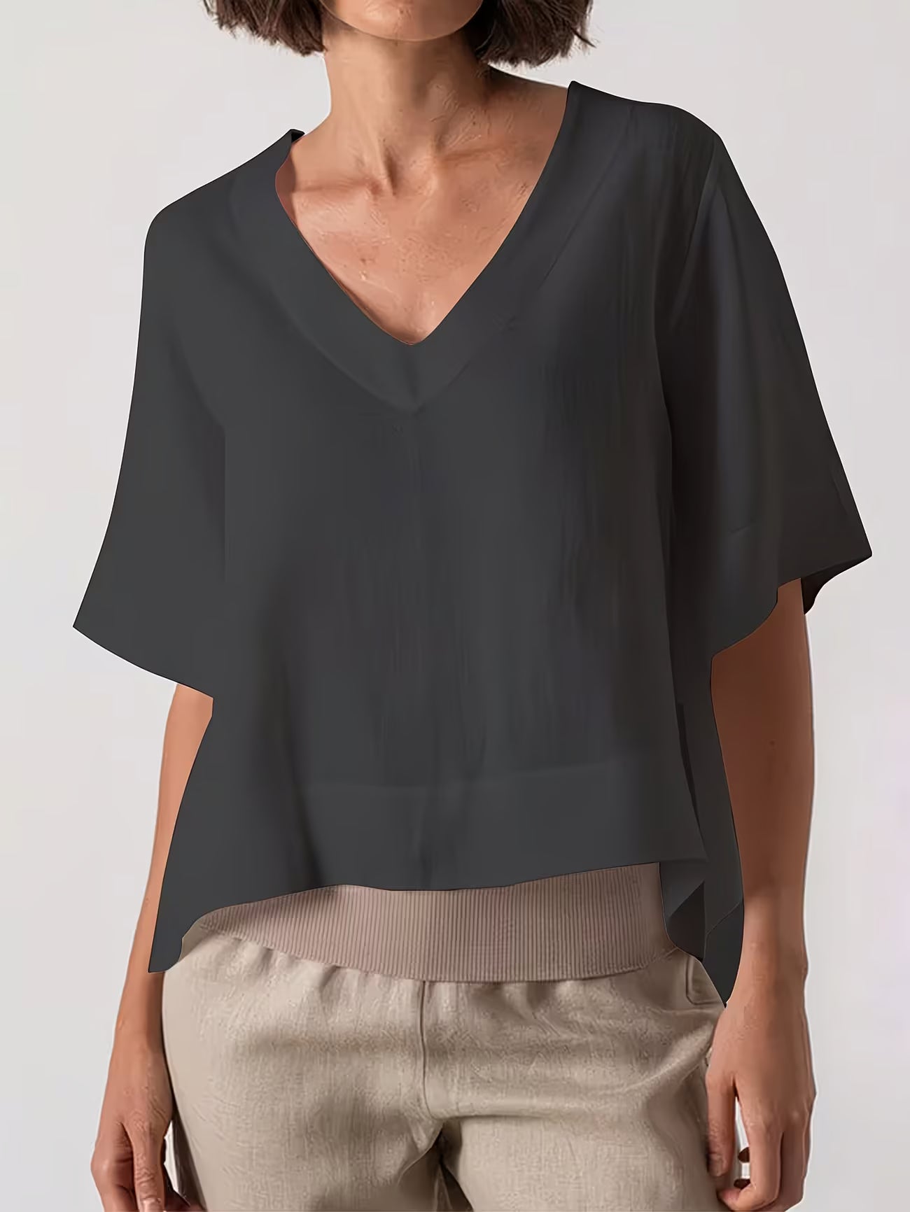 A woman wearing a dark charcoal gray V-neck blouse with short wide sleeves and light beige pants, shown from the neck down against a light gray studio background.