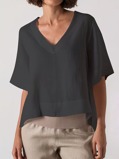 A woman wearing a dark charcoal gray V-neck blouse with short wide sleeves and light beige pants, shown from the neck down against a light gray studio background.